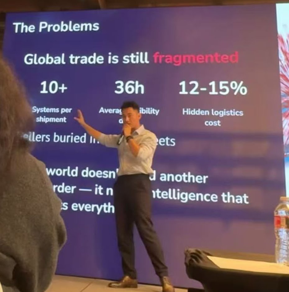Jay Li on stage with a microphone, pointing at a slide titled The Problems: Global trade is still fragmented